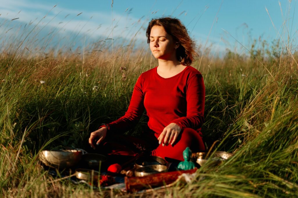 Serene scene of a woman meditating on grass with singing bowls under a blue sky.